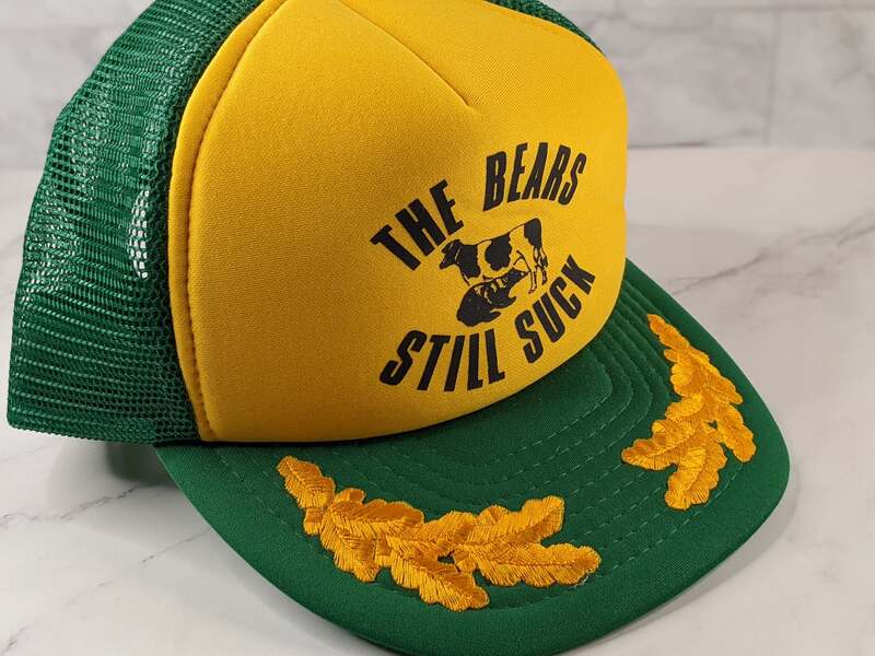A yellow and green baseball cap that features an image of a cow with the words "The Bears Still Suck"